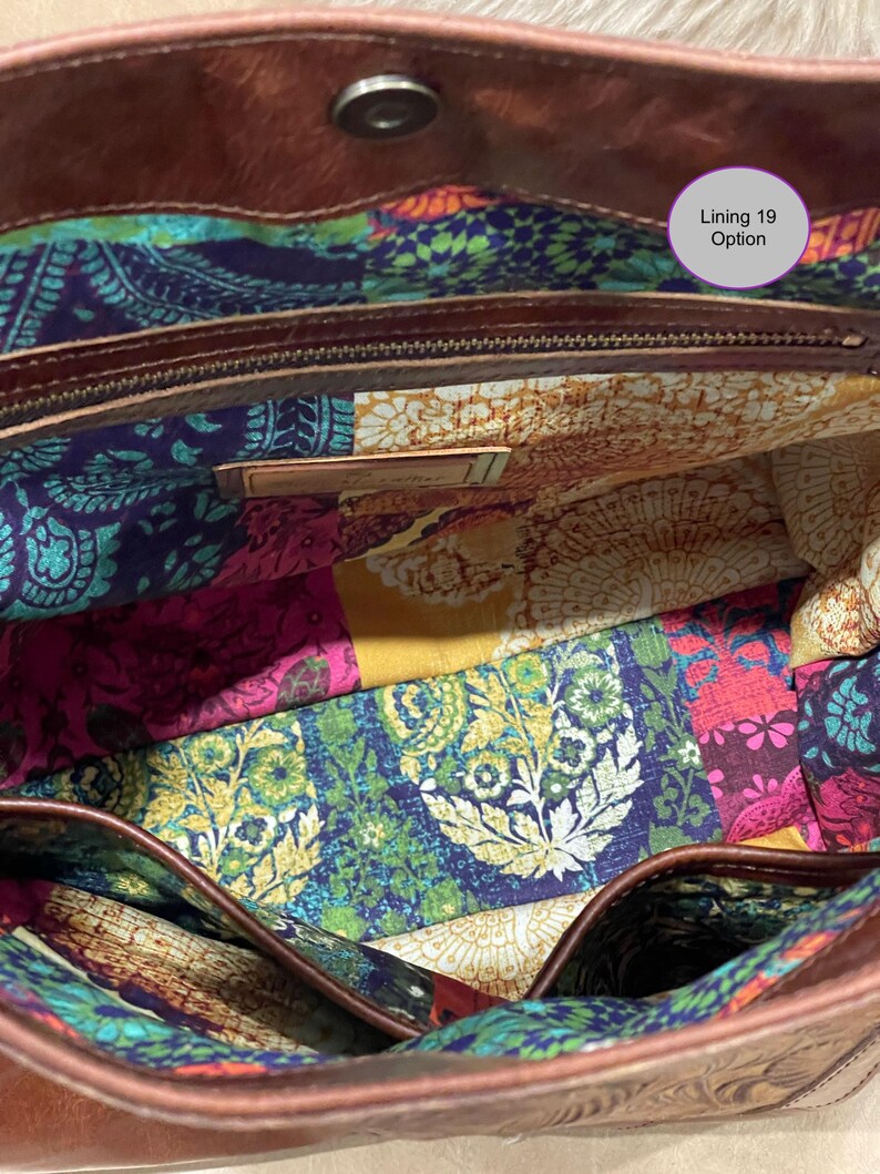 May include: A brown leather bag interior featuring a colorful fabric lining with a patchwork pattern of paisley and floral motifs in shades of blue, yellow, pink, and green. A metal zipper pocket and a small rectangular metal tag are visible.