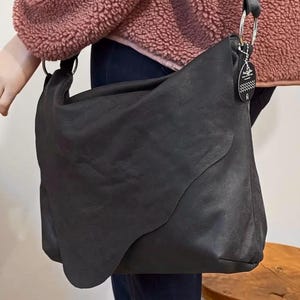 May include: A large, black leather shoulder bag with a unique, curved design. The bag features a long strap with a silver ring attachment and a small tag. The bag is held by a person wearing a pink jacket.