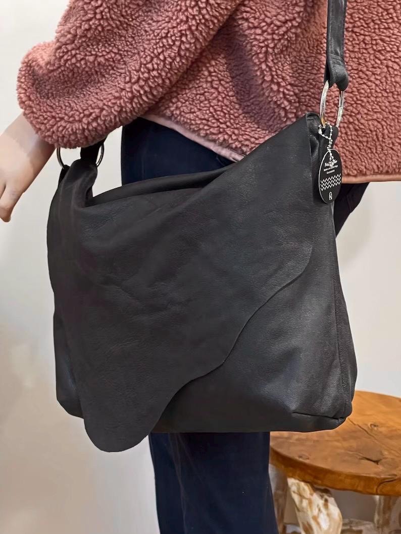 May include: A large, black leather shoulder bag with a unique, curved design. The bag features a long strap with a silver ring attachment and a small tag. The bag is held by a person wearing a pink jacket.