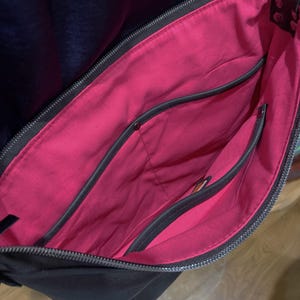 May include: Black shoulder bag with a bright pink interior. The bag has multiple pockets and a zipper closure. The interior lining is a vibrant pink, contrasting with the black exterior. The bag appears to be made of leather or a similar material.