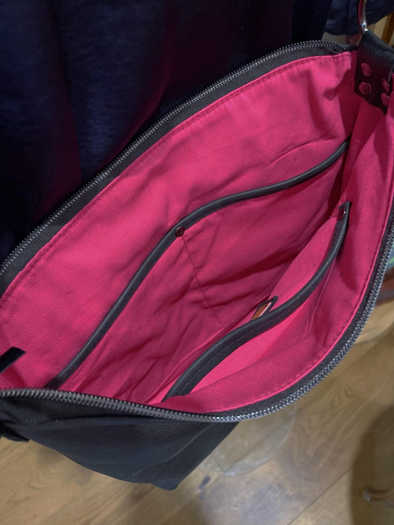 May include: Black shoulder bag with a bright pink interior. The bag has multiple pockets and a zipper closure. The interior lining is a vibrant pink, contrasting with the black exterior. The bag appears to be made of leather or a similar material.