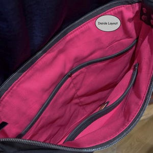 May include: A black bag with a vibrant pink interior lining. The bag features a top zipper closure and multiple internal pockets with dark gray trim. A small oval label with the text Inside Layout is visible near the top of the pink fabric.