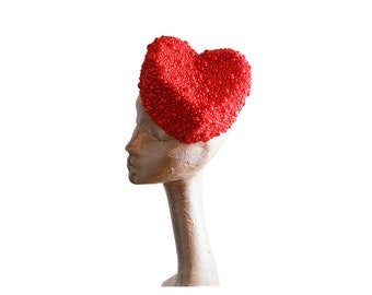May include: A red heart-shaped hat made of small beads. The hat is worn on a mannequin's head.