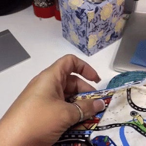 May include: A person's hand is applying white glue to a colorful fabric with a road and car design. The fabric is being used to create a craft project.