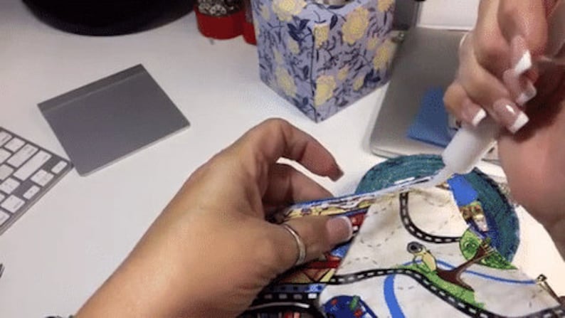 May include: A person's hand is applying white glue to a colorful fabric with a road and car design. The fabric is being used to create a craft project.