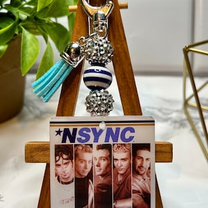 May include: A silver keychain with a blue and white striped bead, a silver rhinestone bead, a silver tassel, and a clear plastic charm with a picture of the boy band *NSYNC.