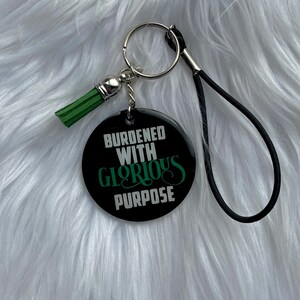 May include: Black and green keychain with a round button that says "burdened with glorious purpose". The keychain has a black strap and a green tassel.