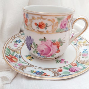 May include: A white porcelain teacup and saucer with a floral pattern in shades of pink, purple, orange, yellow, and green. The cup and saucer have a gold trim.