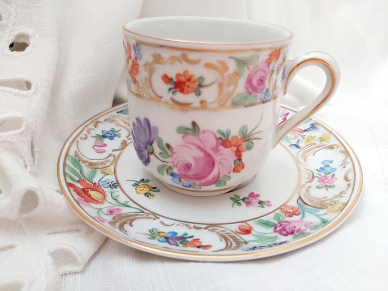 May include: A white porcelain teacup and saucer with a floral pattern in shades of pink, purple, orange, yellow, and green. The cup and saucer have a gold trim.