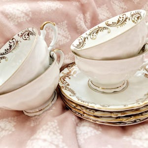 Könnte beinhalten: Eine Sammlung weißer Teetassen und Untertassen mit goldenen Verzierungen und dekorativen Mustern. Die Teetassen haben geschwungene Henkel und stehen auf passenden Untertassen. Das Set ist auf einer rosa Oberfläche mit floralem Muster angeordnet.