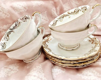 May include: A collection of white teacups and saucers with gold trim and decorative patterns. The teacups have curved handles and sit atop matching saucers. The set is arranged on a pink floral patterned surface.