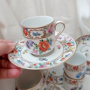 May include: A set of vintage porcelain teacups and saucers with floral patterns in shades of pink, blue, and yellow. The cups and saucers have gold trim and are decorated with delicate floral designs.