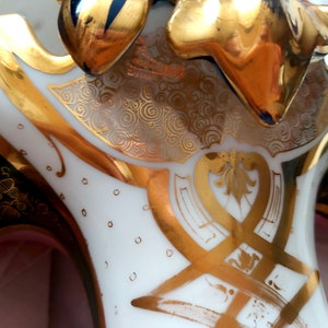 K&ouml;nnte beinhalten: Eine wei&szlig;e Porzellanvase mit goldenen und gr&uuml;nen Akzenten. Die Vase hat eine einzigartige Form mit drei Ausg&uuml;ssen und ein dekoratives Design aus Wirbeln und floralen Mustern.