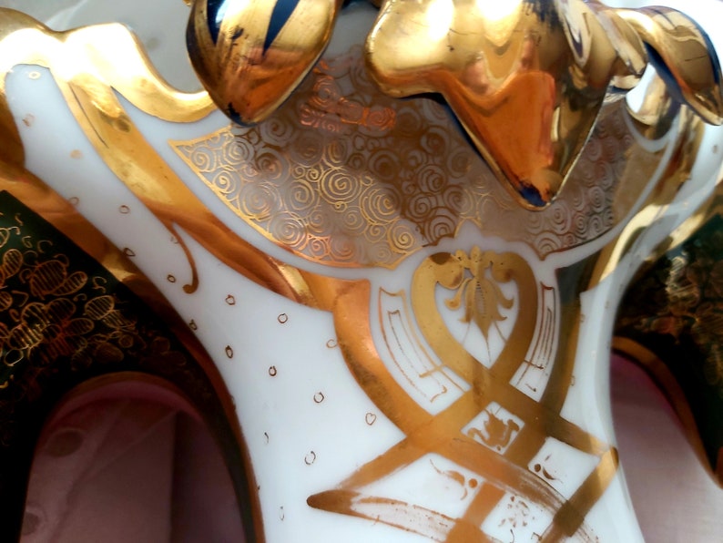 K&ouml;nnte beinhalten: Eine wei&szlig;e Porzellanvase mit goldenen und gr&uuml;nen Akzenten. Die Vase hat eine einzigartige Form mit drei Ausg&uuml;ssen und ein dekoratives Design aus Wirbeln und floralen Mustern.
