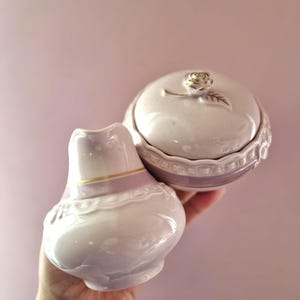 Può includere: Un vaso in ceramica bianca e un piccolo contenitore con coperchio, entrambi con un accento lavanda. Il vaso ha una forma bulbosa con una fascia dorata, mentre il contenitore ha un dettaglio di rosa dorata sul coperchio. Su sfondo rosa.