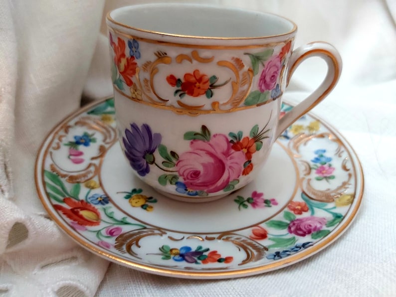 May include: A white porcelain teacup and saucer with a gold rim and floral pattern. The cup has a handle and the saucer has a scalloped edge.