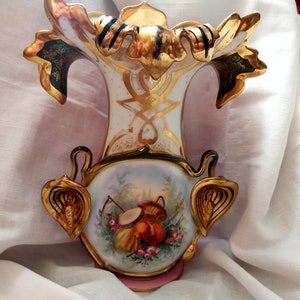 K&ouml;nnte beinhalten: Eine wei&szlig;e Porzellanvase mit goldenen und gr&uuml;nen Akzenten. Die Vase hat eine breite &Ouml;ffnung und einen schmalen Hals und ist mit einem Blumendesign verziert. Die Vase steht auf einer wei&szlig;en Stoffoberfl&auml;che.