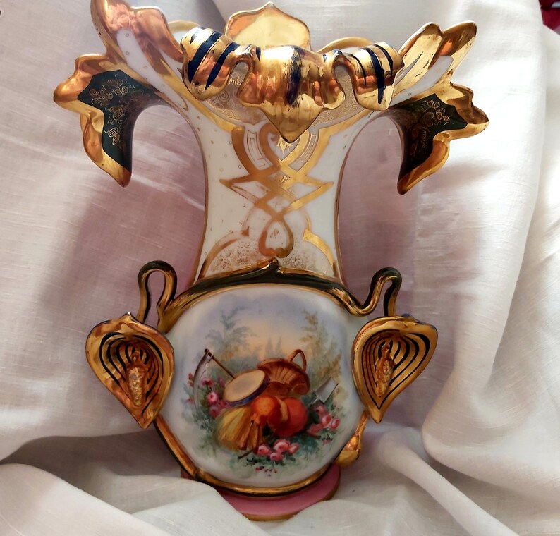 K&ouml;nnte beinhalten: Eine wei&szlig;e Porzellanvase mit goldenen und gr&uuml;nen Akzenten. Die Vase hat eine breite &Ouml;ffnung und einen schmalen Hals und ist mit einem Blumendesign verziert. Die Vase steht auf einer wei&szlig;en Stoffoberfl&auml;che.
