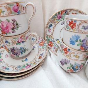 May include: A set of vintage porcelain teacups and saucers with floral patterns and gold accents. The cups and saucers are stacked and arranged on a white tablecloth.