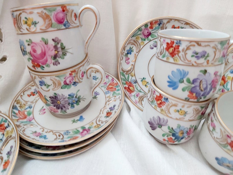 May include: A set of vintage porcelain teacups and saucers with floral patterns and gold accents. The cups and saucers are stacked and arranged on a white tablecloth.