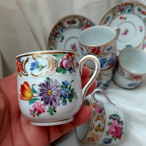 May include: Set of five porcelain teacups and saucers with floral patterns in shades of pink, blue, yellow, and green. The cups and saucers have gold trim.