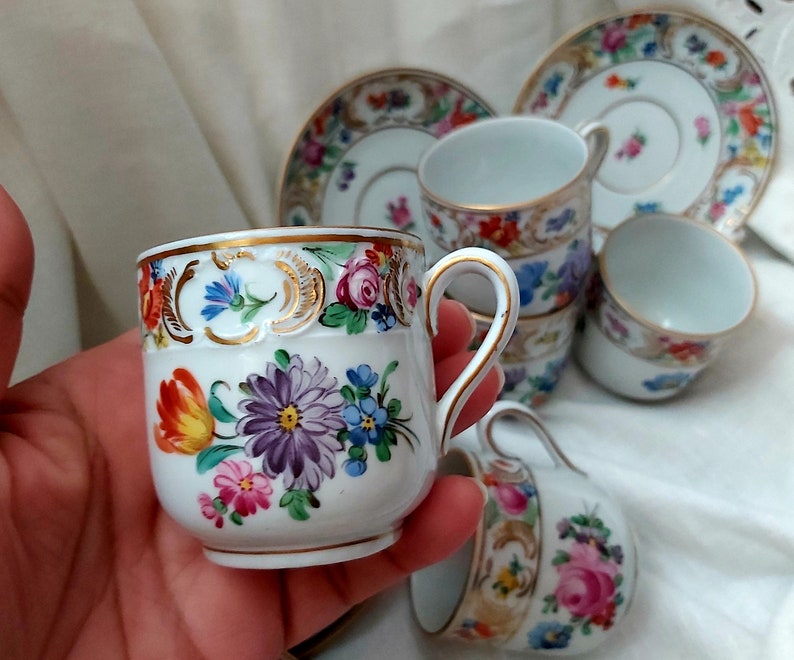May include: Set of five porcelain teacups and saucers with floral patterns in shades of pink, blue, yellow, and green. The cups and saucers have gold trim.