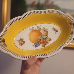 Puede incluir: Plato de servir ovalado de porcelana amarilla y blanca con borde festoneado. El plato presenta un diseño central de frutas con una naranja, bayas y otras frutas. El plato tiene dos asas pequeñas y un borde calado.
