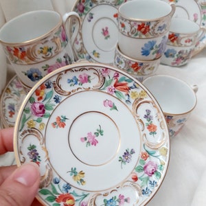 May include: A set of vintage porcelain teacups and saucers with floral patterns and gold accents. The cups and saucers are white with a delicate floral design in shades of pink, blue, yellow, and green. The gold accents are around the rim of the cups and saucers, and there is a gold band around the center of the cups.