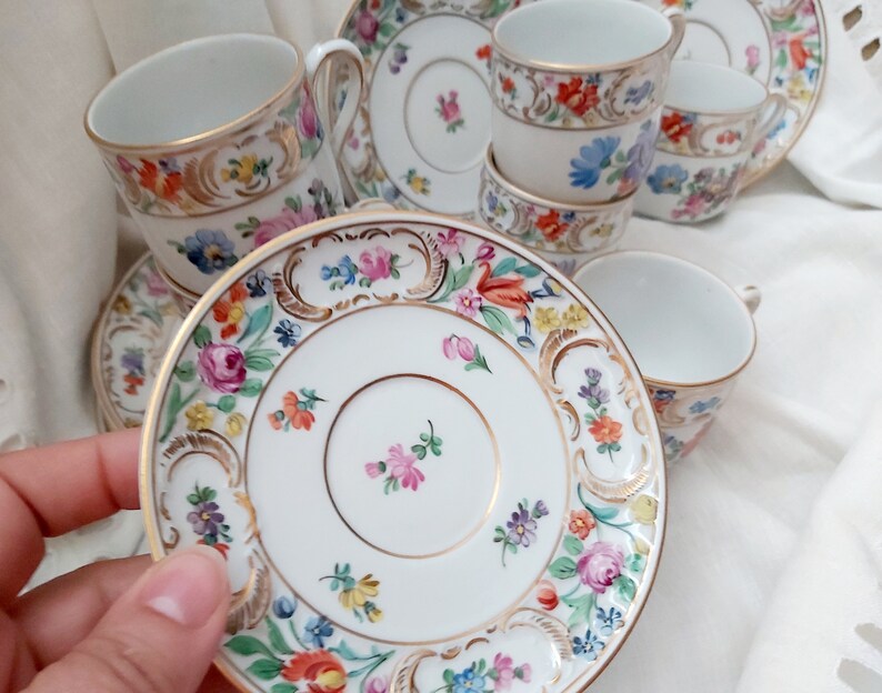 May include: A set of vintage porcelain teacups and saucers with floral patterns and gold accents. The cups and saucers are white with a delicate floral design in shades of pink, blue, yellow, and green. The gold accents are around the rim of the cups and saucers, and there is a gold band around the center of the cups.