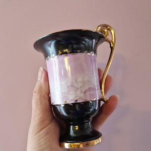 May include: A vintage teacup with a black rim and base, a light purple center, and a gold handle. The cup has gold trim around the rim and base. The cup is held in a hand against a pink background.