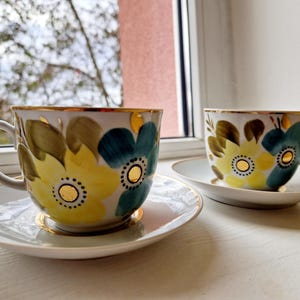 May include: Two white teacups and saucers with floral designs. The cups and saucers have a gold rim and are decorated with yellow, blue, and green flowers. The teacups are on a white surface.