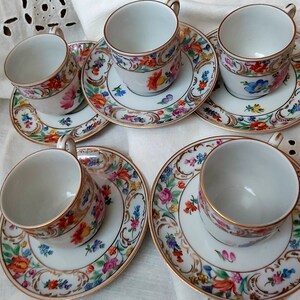 May include: Set of five white porcelain teacups and saucers with gold trim and floral designs. The cups and saucers feature a variety of colourful flowers and leaves.