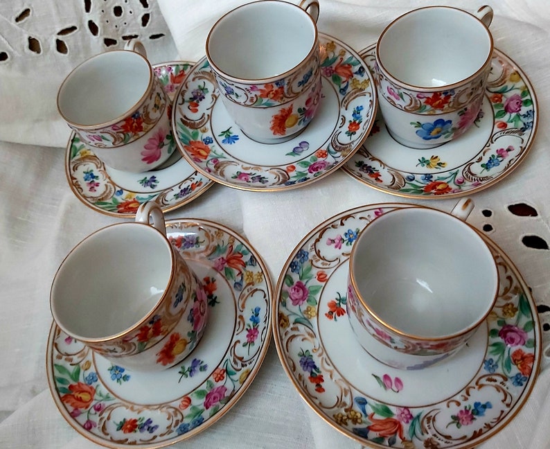 May include: Set of five white porcelain teacups and saucers with gold trim and floral designs. The cups and saucers feature a variety of colourful flowers and leaves.
