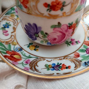 May include: A white porcelain teacup and saucer with a gold trim and floral pattern. The flowers are painted in shades of pink, red, blue, yellow, and green.