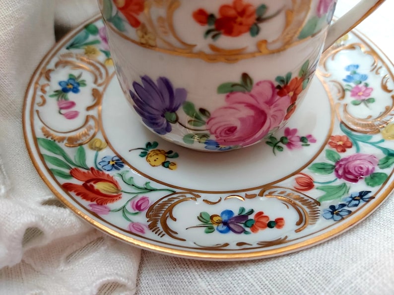 May include: A white porcelain teacup and saucer with a gold trim and floral pattern. The flowers are painted in shades of pink, red, blue, yellow, and green.