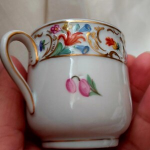 May include: A white porcelain teacup with a gold rim and floral pattern. The cup has a small pink flower painted on the front.