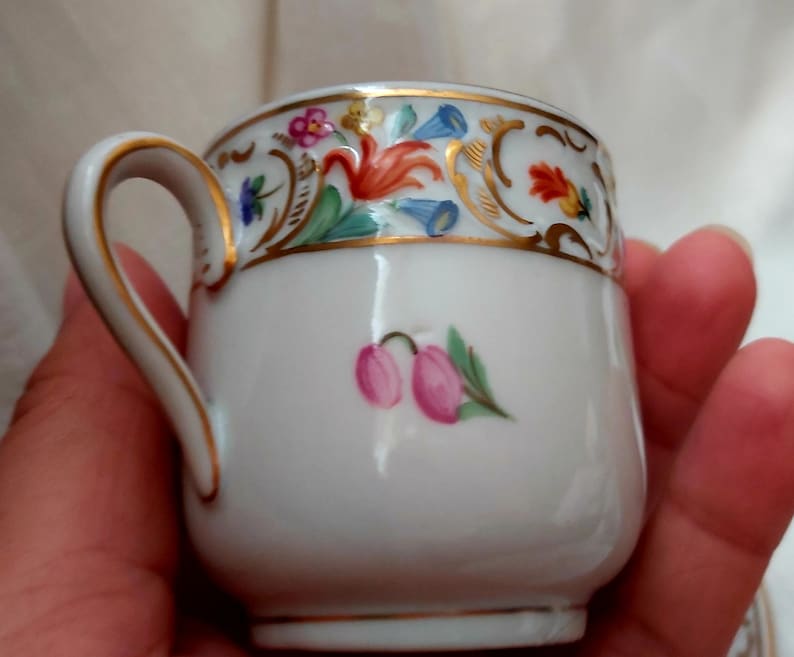 May include: A white porcelain teacup with a gold rim and floral pattern. The cup has a small pink flower painted on the front.