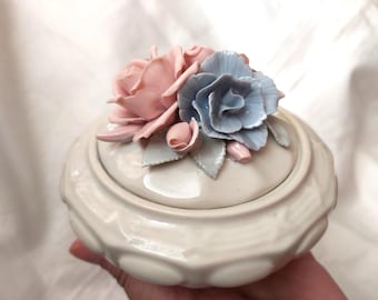Puede incluir: Un joyero redondo de cerámica color blanco hueso con una tapa que presenta decoraciones florales esculpidas en tres dimensiones. La tapa está adornada con un grupo de rosas de cerámica en rosa claro y azul pálido con pequeñas hojas verdes.