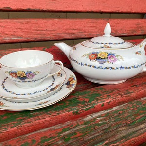 May include: A white teapot, teacup and saucer set with a floral pattern in shades of orange, yellow, pink and green. The teapot has a lid and a spout. The teacup and saucer have a gold rim.