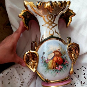 K&ouml;nnte beinhalten: Eine wei&szlig;e Porzellanvase mit goldenen und gr&uuml;nen Akzenten. Die Vase hat ein Blumenmuster und einen Korb mit Fr&uuml;chten auf der Vorderseite. Die Vase ist mit Blattgold verziert und hat einen rosafarbenen Sockel.
