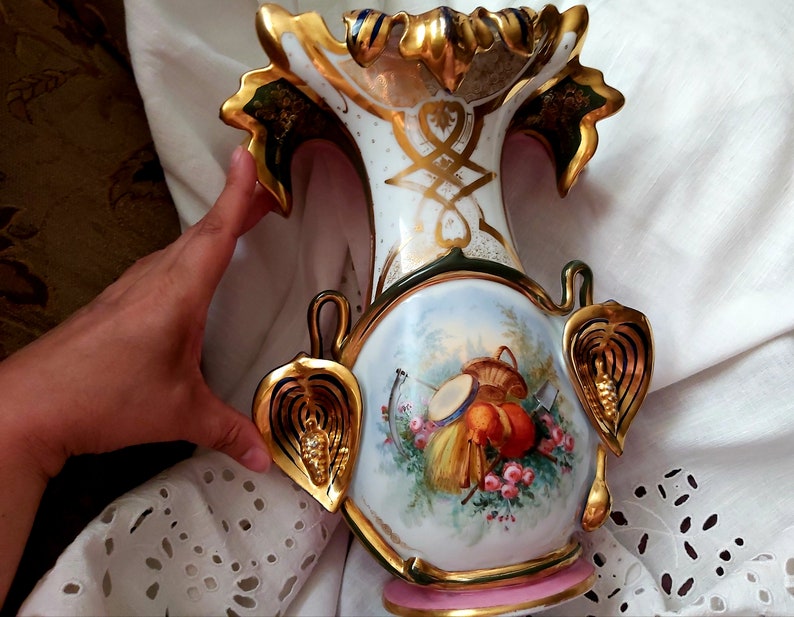 K&ouml;nnte beinhalten: Eine wei&szlig;e Porzellanvase mit goldenen und gr&uuml;nen Akzenten. Die Vase hat ein Blumenmuster und einen Korb mit Fr&uuml;chten auf der Vorderseite. Die Vase ist mit Blattgold verziert und hat einen rosafarbenen Sockel.