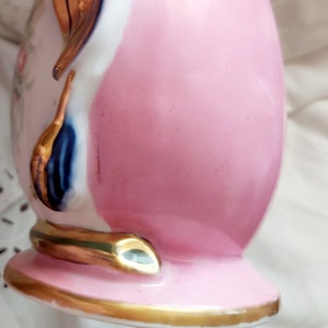 K&ouml;nnte beinhalten: Eine rosa und wei&szlig;e Porzellanvase mit goldenen Akzenten und einem blauen und goldenen Blumendesign. Die Vase hat einen runden K&ouml;rper und einen ausgestellten Boden.