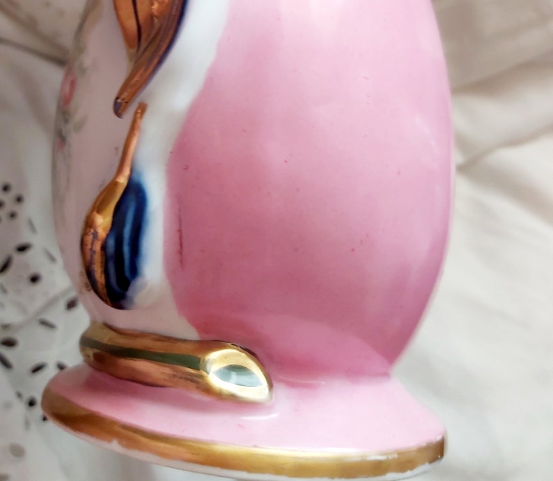 K&ouml;nnte beinhalten: Eine rosa und wei&szlig;e Porzellanvase mit goldenen Akzenten und einem blauen und goldenen Blumendesign. Die Vase hat einen runden K&ouml;rper und einen ausgestellten Boden.