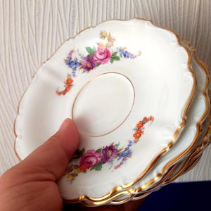 May include: A stack of white porcelain saucers with scalloped edges and gold trim. Each saucer features a delicate floral pattern with pink roses, purple flowers, and small orange blossoms arranged in a curved design around the centre.