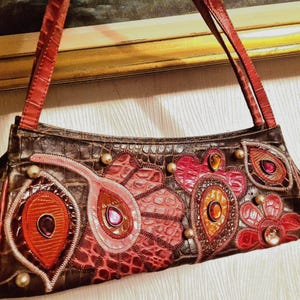 Può includere: Una borsa rettangolare con texture goffrata effetto coccodrillo marrone scuro, dotata di manici rossi effetto coccodrillo e applicazioni decorative floreali e a forma di paisley nei toni del rosso e dell'arancione, arricchita da gemme e perline.
