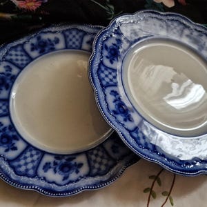 May include: Two antique white plates with a blue floral and geometric design around the rim. The plates have a scalloped edge and a cream-coloured centre. The plates are likely made of porcelain or ceramic.
