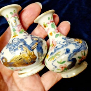 Op de afbeelding: Twee kleine, antieke porseleinen vazen. De vazen zijn wit met blauwe, gouden en roze bloemen- en figuratieve ontwerpen. De vazen hebben een bolvorm met een smalle hals en uitlopende rand. De bases zijn licht uitlopend en hebben een gouden rand.