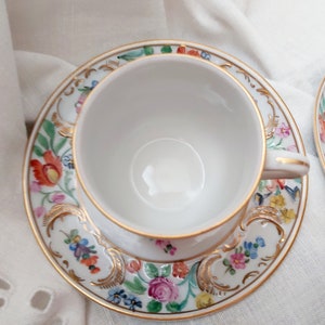 May include: A white teacup and saucer with a floral pattern in shades of pink, yellow, blue, and green. The cup and saucer have a gold trim.
