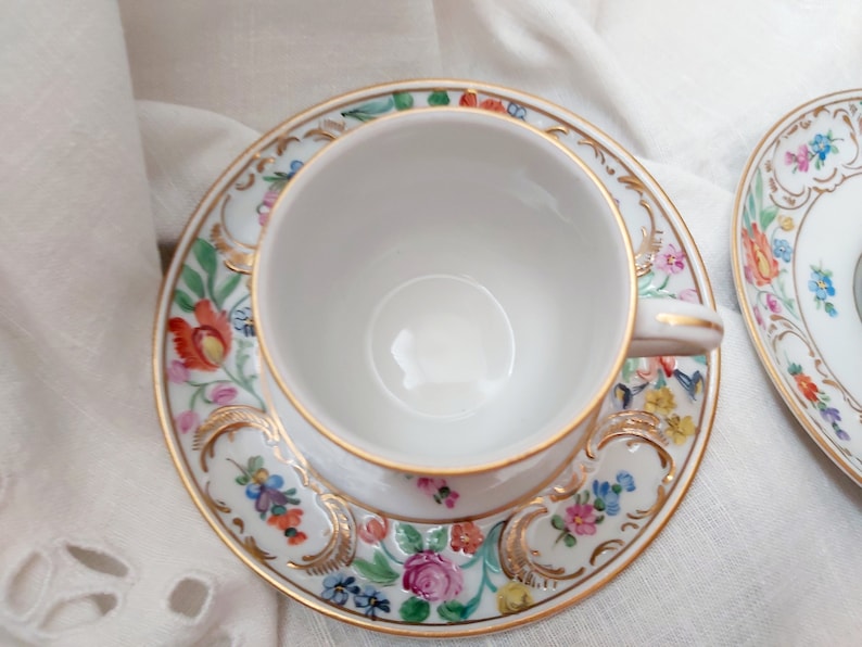 May include: A white teacup and saucer with a floral pattern in shades of pink, yellow, blue, and green. The cup and saucer have a gold trim.