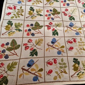 May include: A cream-coloured fabric runner featuring a grid pattern with cross-stitched botanical illustrations in each square. The designs depict various berries, leaves, and twigs in shades of red, blue, green, and yellow on a textured weave.